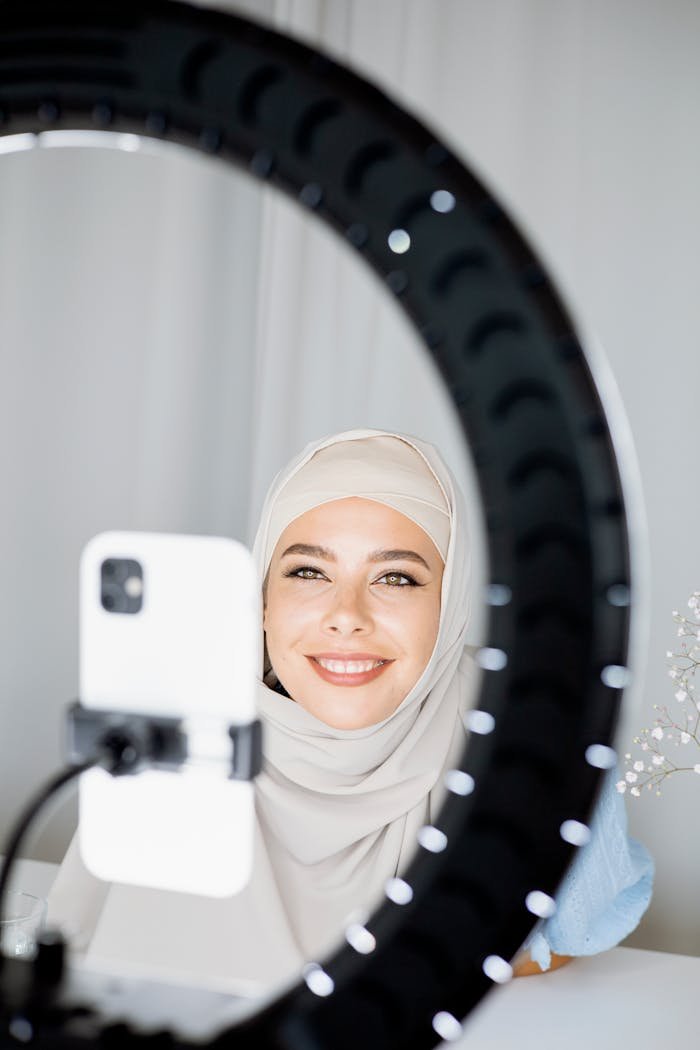 Smiling Muslim woman using a smartphone and ring light for vlogging indoors.